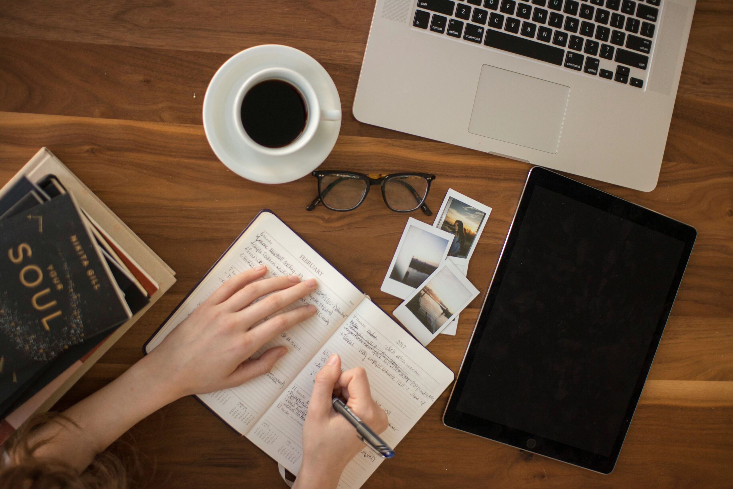 person writing in notebook with glasses, pictures, books, and a laptop on a table.